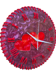 A round, decorative wall clock with a deep purple with swirling resin background. The clock face features red Roman numerals marking the hours, each connected to the edge of the clock by a raised, textured red line that resembles stitching. In the center of the clock face, two overlapping, translucent red hearts are positioned. The clock has simple, white hour and minute hands. The overall aesthetic is handcrafted and romantic.
