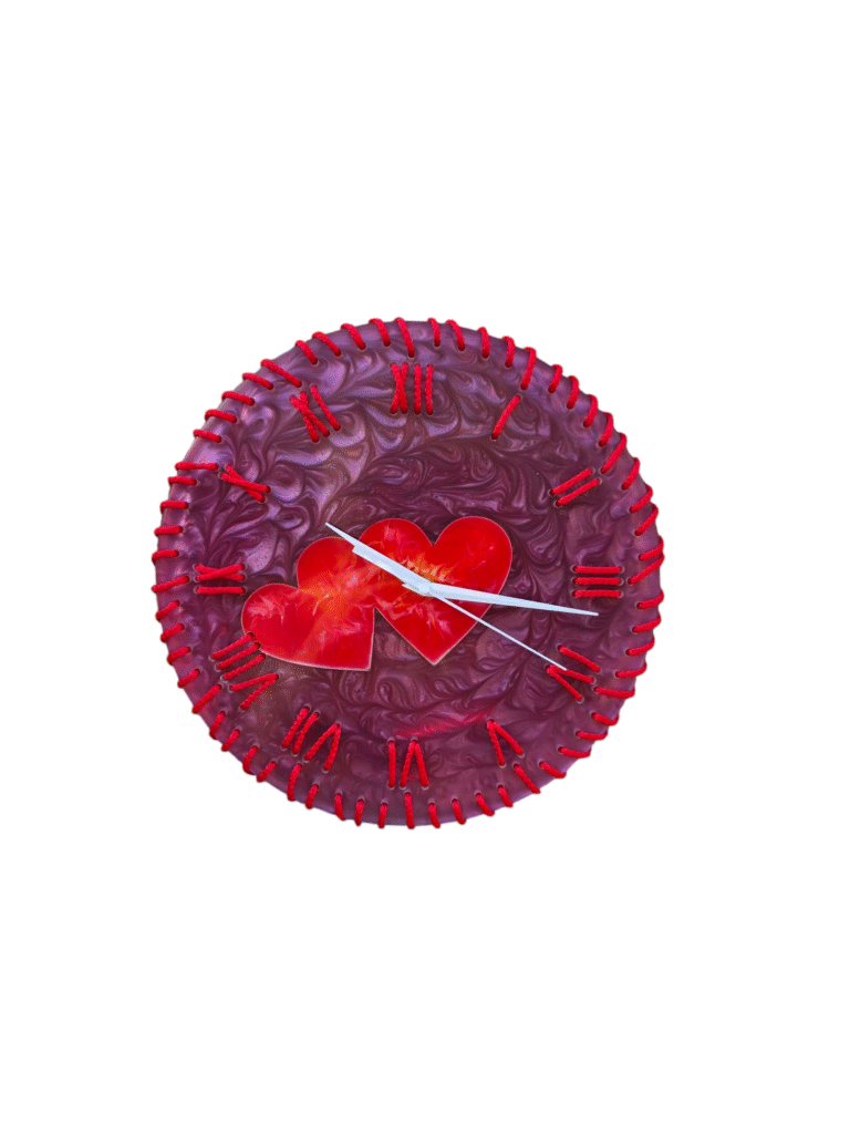 A round, decorative wall clock with a deep purple with swirling resin background. The clock face features red Roman numerals marking the hours, each connected to the edge of the clock by a raised, textured red line that resembles stitching. In the center of the clock face, two overlapping, translucent red hearts are positioned. The clock has simple, white hour and minute hands. The overall aesthetic is handcrafted and romantic.