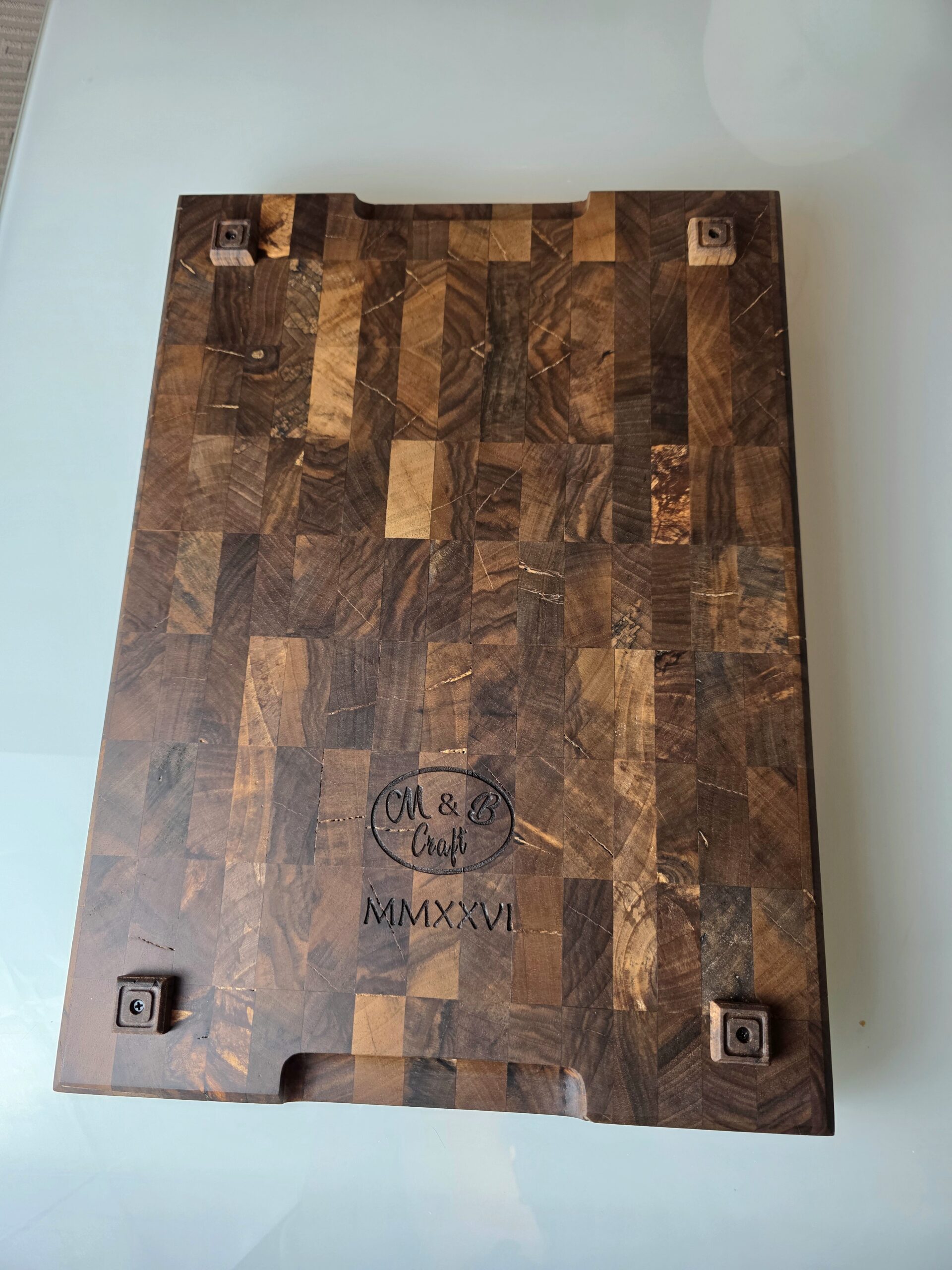 20260120_171146 - M & B CRAFT Underside of walnut cutting board showing custom-crafted solid walnut feet for elevation. Laser engraved M & B Craft logo and MMXXVI date mark. Shows natural wood grain detail.