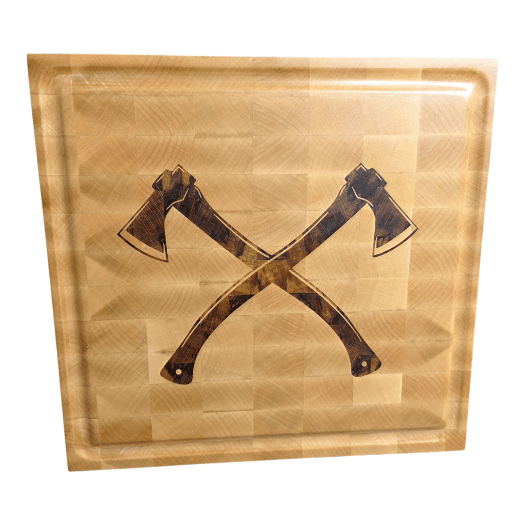 Large end-grain maple butcher block with a custom dark wood double axe inlay and a deep juice groove, sitting on a workshop bench.