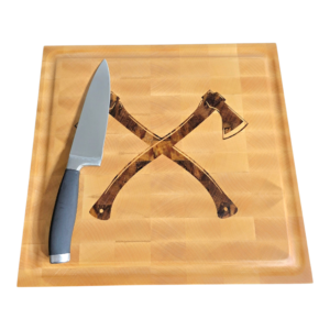 Large end-grain maple butcher block with a custom dark wood double axe inlay and a deep juice groove, sitting on a workshop bench.