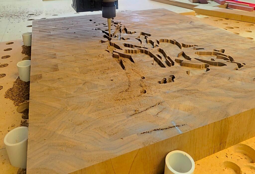 CNC router bit carving curved grooves into a large plywood sheet on a workshop table, with sawdust and white pipes nearby.