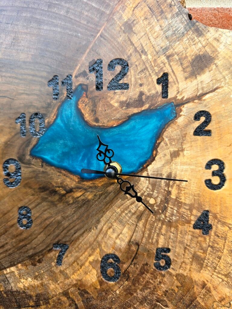 20260728_112232 - M & B CRAFT Close-up of a wooden clock face with carved black numbers and blue epoxy inlays, showing ornate clock hands.